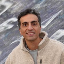 Saber Hafezqorani with a warm smile, in a cream fleece jacket, outdoors in front of a purple-grey painted wall.